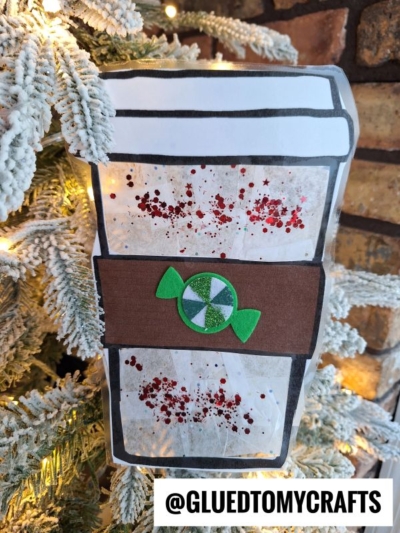 A handmade hot chocolate suncatcher coffee cup ornament with glitter and a green candy decoration hangs on a snow-covered Christmas tree branch, with a “@GLUEDTOMYCRAFTS” label at the bottom right.