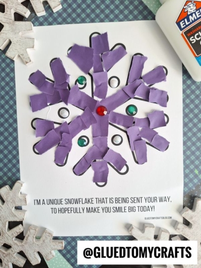 A purple torn paper snowflake collage with green and red gem accents is glued on white paper. The text below reads, “I’M A UNIQUE SNOWFLAKE THAT IS BEING SENT YOUR WAY, TO HOPEFULLY MAKE YOU SMILE BIG TODAY!”.