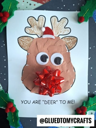 A torn paper reindeer craft featuring large googly eyes, a red gift bow nose, and a tiny Santa hat, is displayed on a sheet that reads YOU ARE DEER TO ME!.