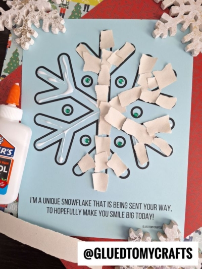 A torn paper snowflake craft with white paper pieces glued onto a blue snowflake outline. A bottle of glue and snowflake decorations are nearby. Text reads: I’m a unique snowflake…to hopefully make you smile big today!.