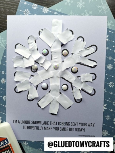 A handmade torn paper snowflake craft on light blue paper, made with white paper strips and round gem stickers, sits on a snowflake-patterned background. Text at the bottom reads: IM A UNIQUE SNOWFLAKE...TO HOPEFULLY MAKE YOU SMILE BIG TODAY!.