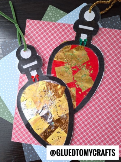 Two handmade Christmas light bulb ornaments with gold foil and glitter decorations, placed on patterned paper backgrounds. Each ornament captures a festive glow reminiscent of a christmas light suncatcher, with a green and a gold string loop attached for hanging.