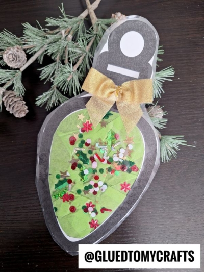 A handmade Christmas light suncatcher craft shaped like a green bulb, decorated with sequins, glitter, and confetti, topped with a gold bow. Pine branches are in the background. @GLUEDTOMYCRAFTS is written at the bottom.