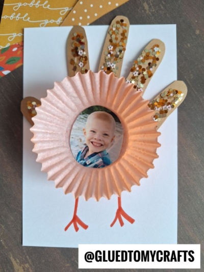 Handprint turkey craft made from brown paper, glitter, and a cupcake liner for the body to create a charming cupcake liner turkey. A child’s photo is placed in the center on a white background, with red lines depicting turkey legs. @GLUEDTOMYCRAFTS text at the bottom.