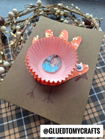 A handmade cupcake liner turkey card features a photo of a child inside an orange cupcake liner, with craft sticks and paper creating the wings and feathers. A decorative wreath and plaid fabric serve as the background.