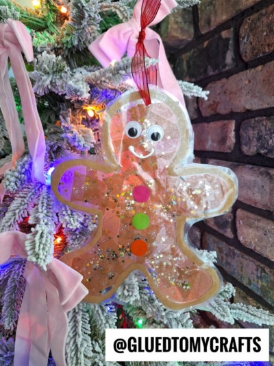 A handmade gingerbread man suncatcher ornament with googly eyes, colored buttons, and glitter hangs on a snowy Christmas tree adorned with pink bows and colorful lights. A label reads @GLUEDTOMYCRAFTS.