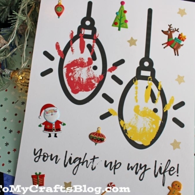 A handmade holiday card features two painted handprints as Christmas light bulbs, one red and one yellow, with festive stickers of Santa, a reindeer, ornaments, and a Christmas tree. The card reads, You light up my life!.
