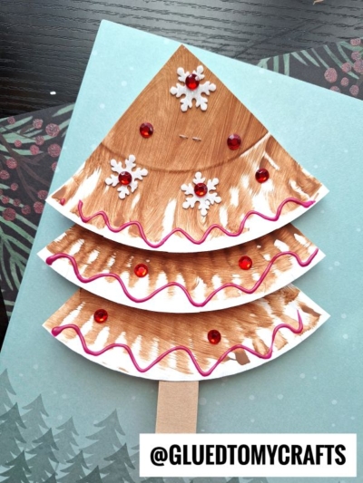 A handmade paper plate gingerbread tree craft, painted and decorated with red sequins, white snowflake stickers, and wavy pink lines, rests on a blue background with pine tree illustrations.