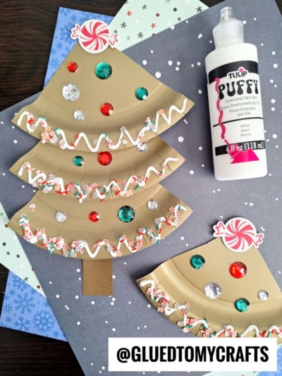 Paper plate Christmas tree crafts, inspired by the look of a paper plate gingerbread tree, are decorated with sequins, faux candy, and puffy paint as they sit on a snowflake-patterned background. A bottle of white puffy paint is nearby. @GLUEDTOMYCRAFTS is written at the bottom.