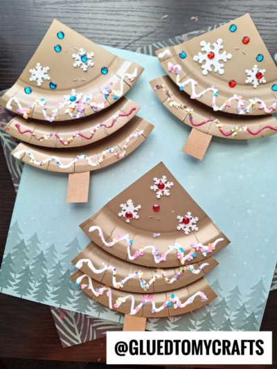 Three paper plate crafts shaped like Christmas trees, including a whimsical paper plate gingerbread tree, decorated with paint, glitter, sequins, and snowflake embellishments, set on winter-themed paper. The text @GLUEDTOMYCRAFTS is at the bottom.