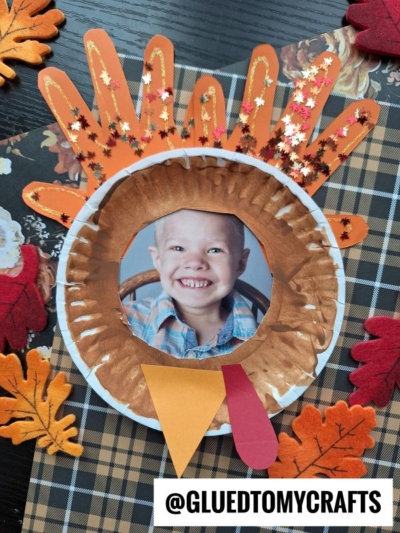 A paper plate turkey frame featuring a child's photo as the turkey's face, with handprint feathers, a paper beak and wattle, all displayed on a plaid background with felt leaves.