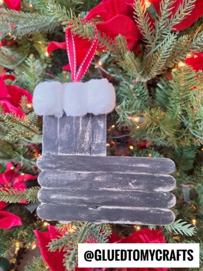 A handmade santa boot ornament shaped like Santa’s black boot, crafted from painted popsicle sticks and white cotton balls, hangs on a decorated Christmas tree with red ribbon and poinsettias. Text reads @GLUEDTOMYCRAFTS.