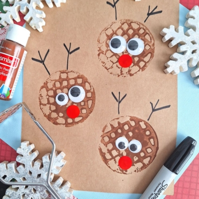 A brown paper features three reindeer faces made from circular brown potato masher stamped craft prints, with googly eyes and red noses. Black lines form antlers. Snowflake decorations, a glue stick, and a black marker surround the paper.