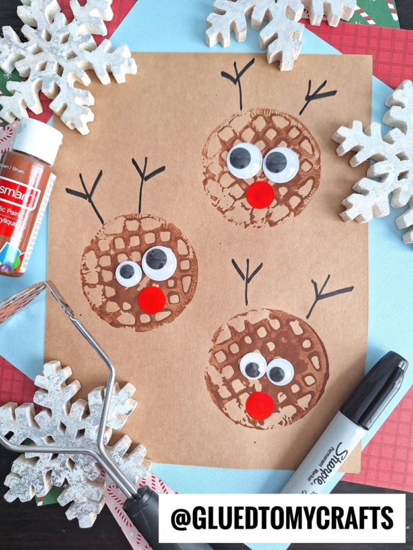 A brown paper features three reindeer faces made from circular brown potato masher stamped craft prints, with googly eyes and red noses. Black lines form antlers. Snowflake decorations, a glue stick, and a black marker surround the paper.