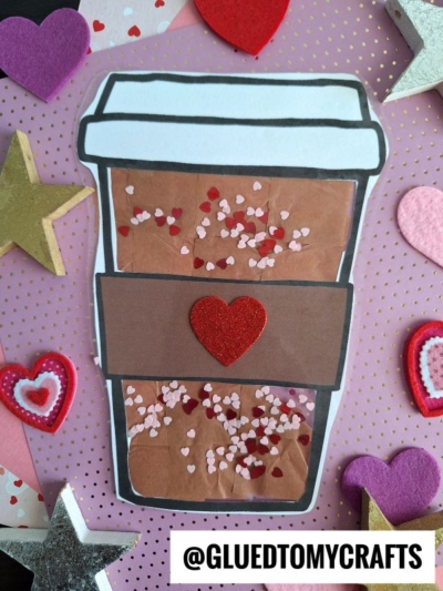 A handmade coffee cup craft with a red heart on a brown sleeve, like a hot chocolate suncatcher, decorated with small heart confetti. Surrounded by colorful felt hearts and stars on a pink polka dot background. Text: @GLUEDTOMYCRAFTS.