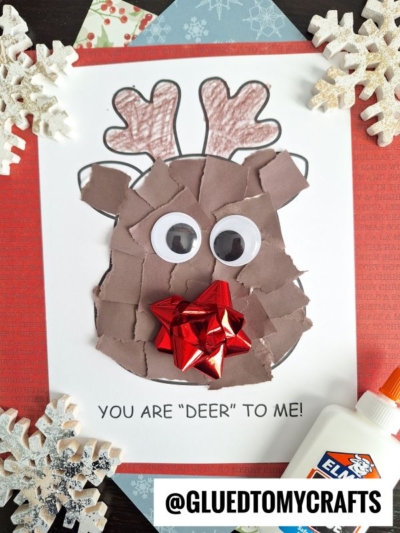 A torn paper reindeer craft features a cute face with brown scraps, googly eyes, a festive red bow nose, and the words YOU ARE DEER TO ME! Snowflake decorations and a glue bottle sit close by.