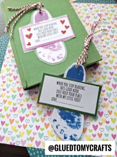 A green book with a footprint bookmark featuring a child's footprint, red heart designs, and a reading quote. Another bookmark with a blue footprint and red-white twine rests on colorful heart-patterned paper. @GLUEDTOMYCRAFTS is visible.