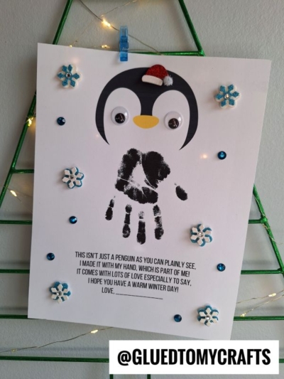 A handmade penguin craft featuring a black handprint body, googly eyes, Santa hat, blue gems, and snowflakes. Perfect for pairing with a handprint penguin poem. Displayed on a wire frame with text and @GLUEDTOMYCRAFTS below.