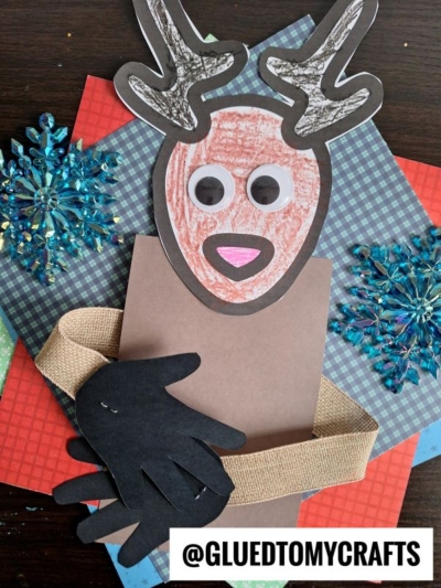 A handmade handprint reindeer hug craft using colored paper, googly eyes, and a burlap ribbon, placed on patterned paper with blue snowflake decorations. The Instagram handle @GLUEDTOMYCRAFTS appears at the bottom.