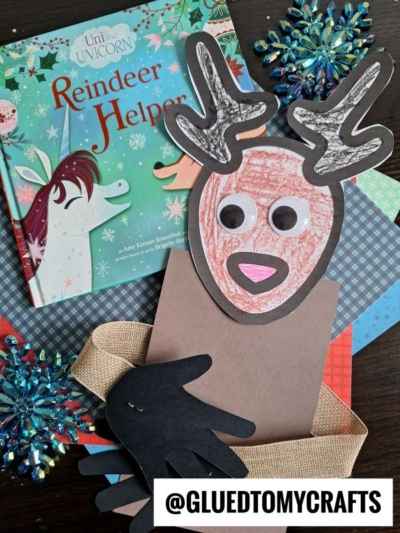 A children’s book titled Uni the Unicorn: Reindeer Helper sits next to a handprint reindeer hug craft with paper antlers and hands, displayed on a table with colorful papers and sparkly snowflake decorations.