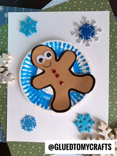 A paper gingerbread man with googly eyes and red buttons, ready for a day of snow tubing, is on a blue, circular background, surrounded by foam and felt snowflakes on a piece of white paper. @GLUEDTOMYCRAFTS is written at the bottom.