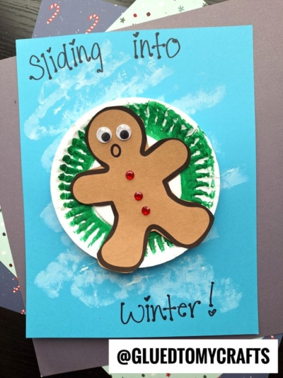 A snow tubing gingerbread man craft with googly eyes and red buttons is glued to a white circle with green paint, resembling a wreath. The blue background says Sliding into winter! and @GLUEDTOMYCRAFTS is shown at the bottom.