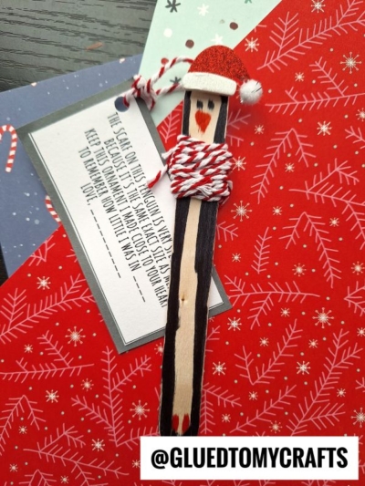 A handmade Santa craft made from a wooden stick with a red hat, drawn face, and red-and-white yarn penguin scarf ornament sits on festive greeting cards. The @GLUEDTOMYCRAFTS handle is visible at the bottom.