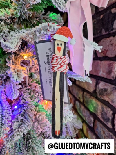 A painted wooden popsicle stick penguin scarf ornament with a red Santa hat hangs on a snowy Christmas tree, next to a pink bow and a brick wall. Text reads “@GLUEDTOMYCRAFTS.”.