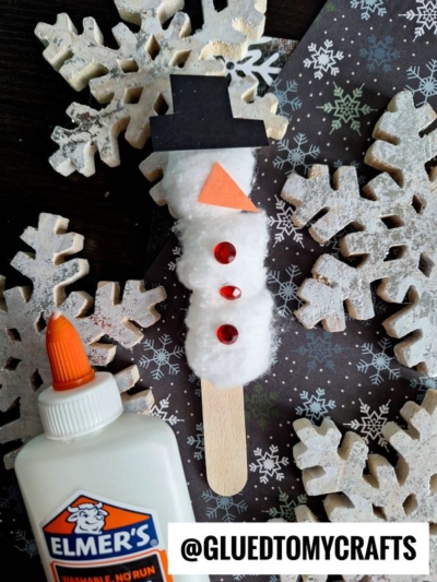 A cotton ball snowman craft features red sequins, an orange paper nose, and a black paper hat on a craft stick, surrounded by wooden snowflakes, Elmer’s glue, and set against a dark snowflake-patterned background.