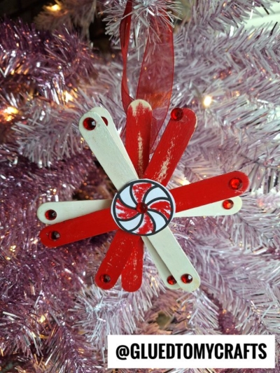 A red and white peppermint candy snowflake ornament, made from popsicle sticks with a swirl center, hangs on a pink Christmas tree. The image includes the text @GLUEDTOMYCRAFTS at the bottom.