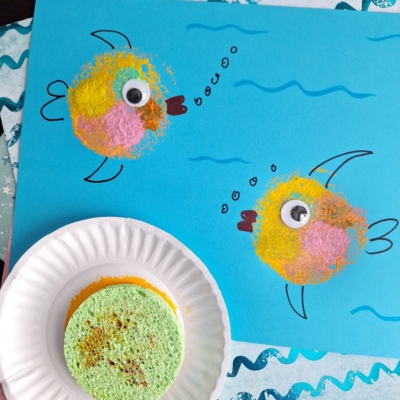 A blue paper displays two fish crafted from painted sponges and googly eyes, with drawn bubbles and waves—perfect for Sponge Painting Ideas for Kids. A paint-dabbed sponge rests on a plate. The text @GLUEDTOMYCRAFTS appears below.