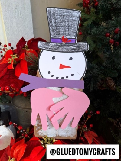 A handmade paper bag snowman craft with a paper hat, face, scarf, and pink handprint cutouts for arms, surrounded by red flowers and greenery. The handle @GLUEDTOMYCRAFTS is displayed at the bottom.