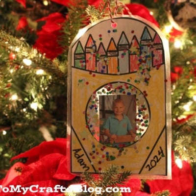A crayon box ornament featuring a circle photo of a child, hand-drawn crayons, and glitter hangs on a Christmas tree with red ribbon.