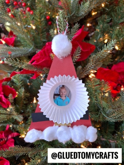 Handmade cupcake liner Santa Christmas tree ornament featuring a smiling child's photo, decorated with cotton balls and hanging among red poinsettias and twinkling lights.