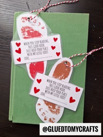 Two footprint bookmarks featuring red and brown child footprints, heart stickers, and a sweet message about holding your place with a little foot are tied with red and white string, resting on a green book. @GLUEDTOMYCRAFTS watermark is visible.