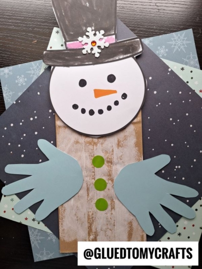 A charming paper bag snowman craft featuring a black hat, white face with a carrot nose, button eyes, and dotted mouth. Blue hand-shaped cutouts form the arms, all set against a background of snowflake-patterned paper.