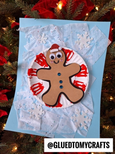 A snow tubing gingerbread man made of brown paper, red paint, googly eyes, blue buttons, and a tiny Santa hat sits on a plate with tissue paper snow and snowflakes, displayed in front of a glowing Christmas tree.