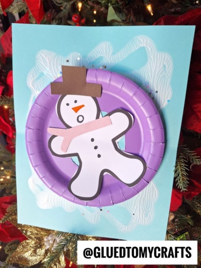 A handmade snow tubing snowman craft using a paper plate, colored paper, and markers is displayed against a blue background with holiday decorations and the Instagram handle @GLUETOMYCRAFTS at the bottom.