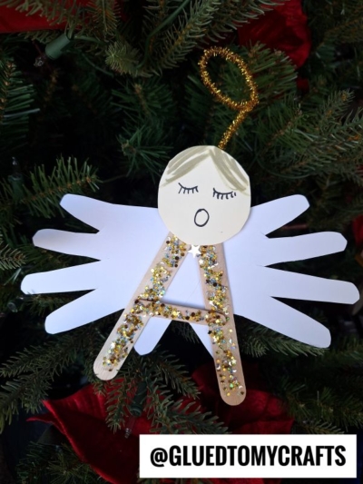 A Christmas ornament shaped like an angel, featuring “A” is for Angel with popsicle sticks and gold glitter, white paper handprint wings, a round face with drawn features, and a gold pipe cleaner halo.