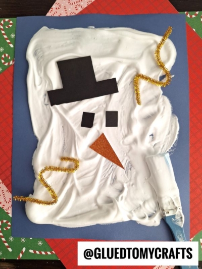 A puffy painted melted snowman face made with white shaving cream, a black paper hat, black square eyes, an orange triangle nose, and gold pipe cleaners on blue paper, placed over red patterned paper. Handle @GLUETOMYCRAFTS is at the bottom.