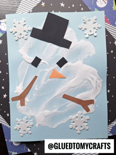 A puffy painted melted snowman made from white paint on blue paper, featuring a black hat, eyes, and mouth, along with an orange nose and arms. White paper snowflakes are scattered around the artwork. The image has the text @GLUEDTOMYCRAFTS at the bottom.