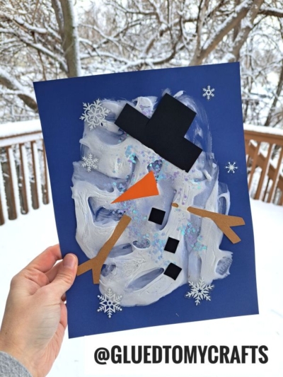 A hand holds a blue sheet with a puffy painted melted snowman, featuring paper shapes for the hat, arms, nose, and buttons, plus snowflake stickers. Snowy trees and a wooden railing are in the background. Text reads @GLUEDTOMYCRAFTS.