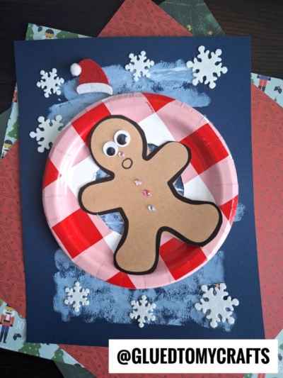 A snow tubing gingerbread man craft made from brown paper and googly eyes sits on a red and white paper plate with snowflake decorations, a Santa hat, and a blue painted background. The text @GLUEDTOMYCRAFTS is at the bottom right.