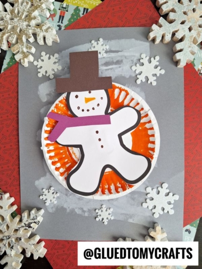 A paper plate snow tubing snowman craft featuring a brown top hat and purple scarf, surrounded by white snowflakes, set on gray paper with an orange background. @GLUEDTOMYCRAFTS is written at the bottom.