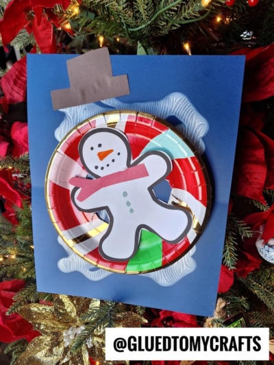 A handmade snow tubing snowman craft features a paper plate with colorful stripes, a paper snowman cutout, and a brown paper hat, set against blue paper and displayed near a Christmas tree. @GLUEDTOMYCRAFTS is at the bottom.
