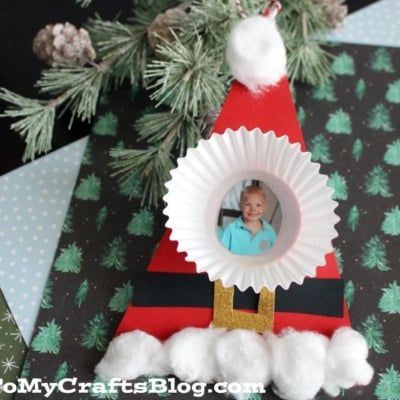 A handmade cupcake liner Santa craft featuring a child's photo as Santa's face, decorated with paper, cotton balls, and displayed on patterned paper with festive winter and tree designs.