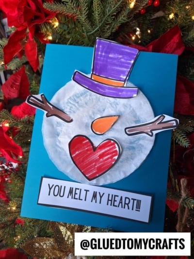 A coffee filter melted snowman craft made from a paper plate with colored paper features, including a purple hat, heart-shaped nose and mouth, and stick arms. A sign reads YOU MELT MY HEART!!! and the craft rests on a Christmas tree.