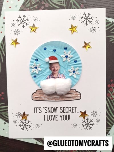 A handmade card features a child’s photo in a cupcake liner snow globe, decorated with cotton, snowflakes, sequins, and stars. The card says: “It’s Snow Secret... I Love You!” @GLUEDTOMYCRAFTS is at the bottom.