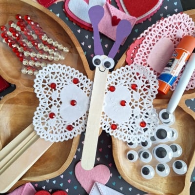 Valentine’s Day butterfly craft made from a popsicle stick, heart doilies, googly eyes, felt hearts, and red gems—perfect for those seeking fun Valentine’s Day craft ideas for kids. Surrounded by extra craft supplies and decorations on a table.