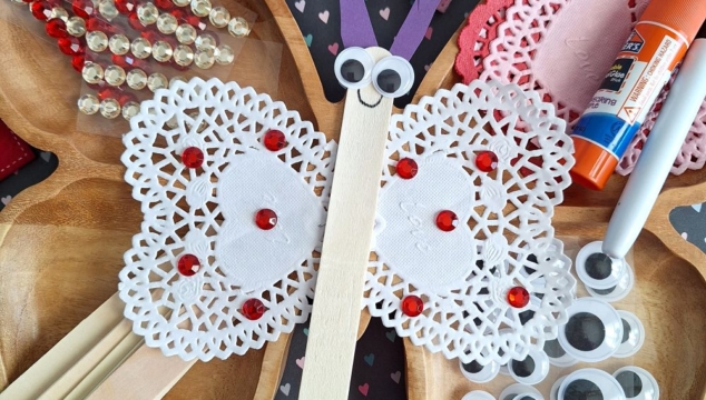 Valentine’s Day butterfly craft made from a popsicle stick, heart doilies, googly eyes, felt hearts, and red gems—perfect for those seeking fun Valentine’s Day craft ideas for kids. Surrounded by extra craft supplies and decorations on a table.